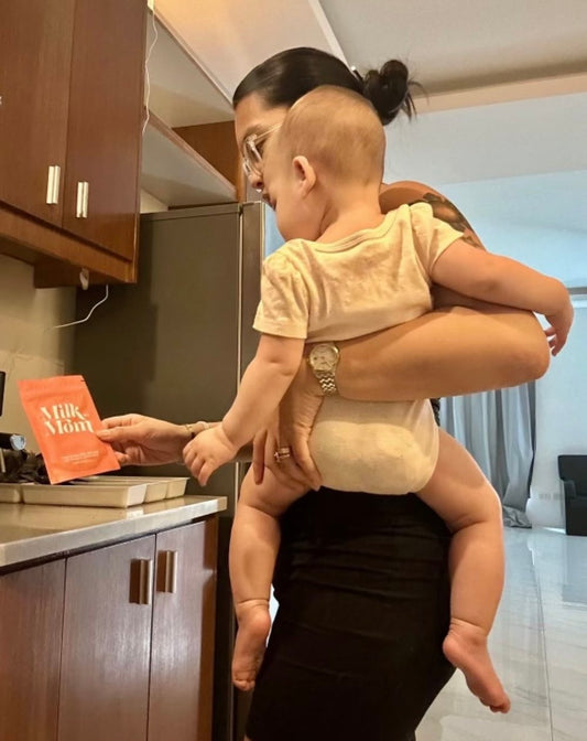 Mother holding baby while preparing milk in kitchen