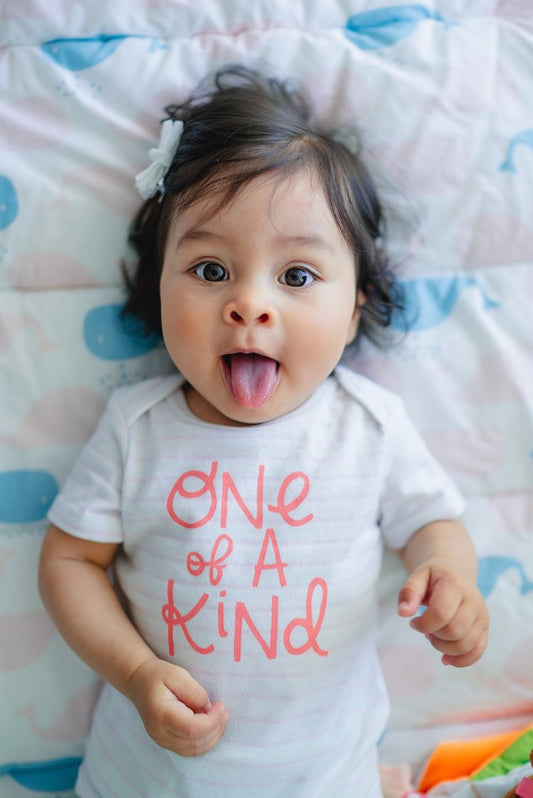 Baby lying on a blanket with whale patterns, sticking out their tongue and wearing a white onesie that says 'One of a Kind' in red letters