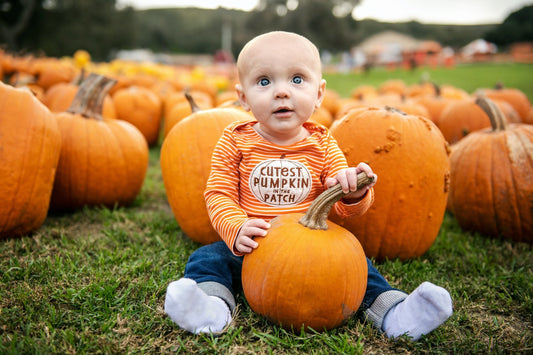 Baby sitting among pumpkins at a pumpkin patch, wearing an orange striped shirt with the words ‘Cutest Pumpkin in the Patch.