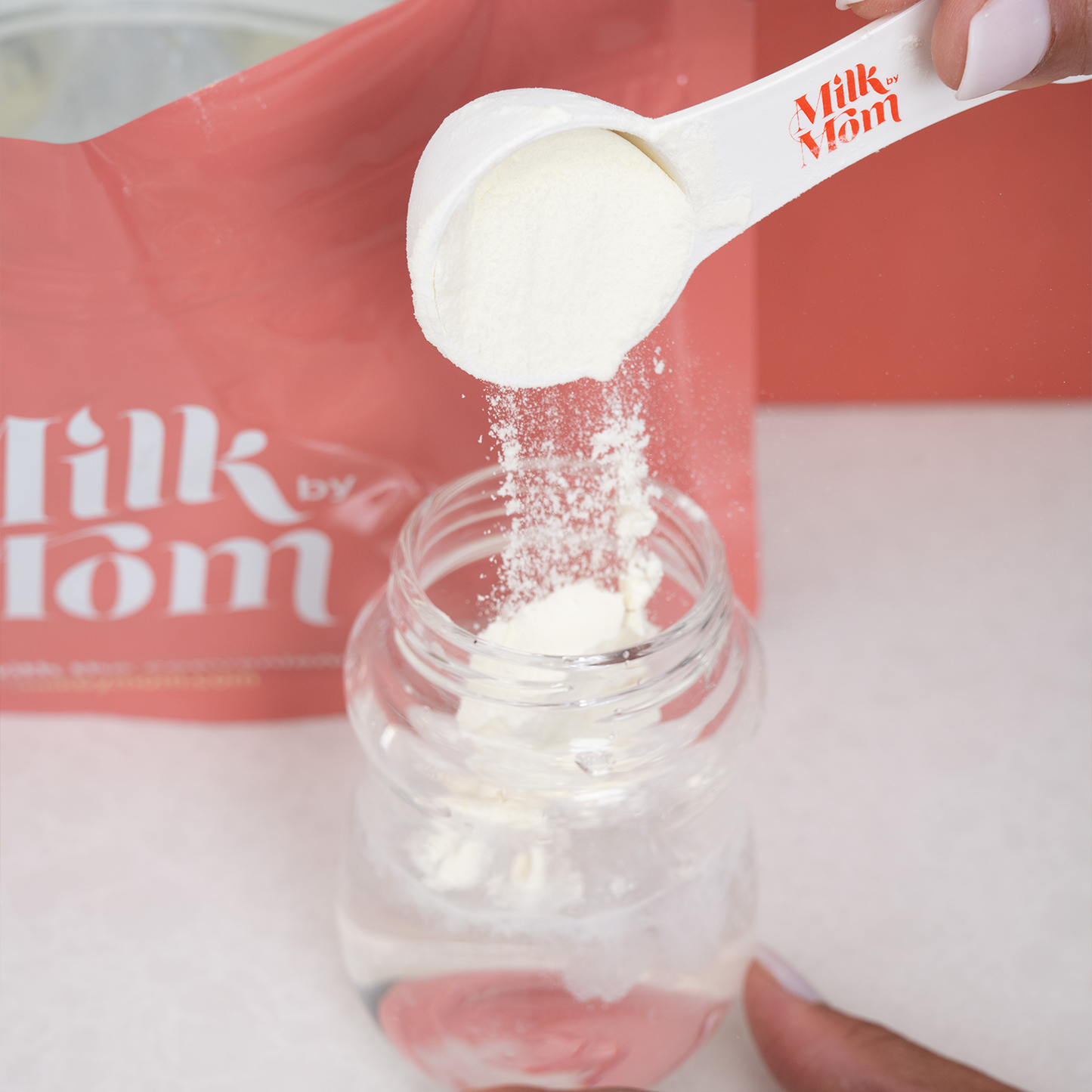 Hand holding a scoop of powdered breast milk, pouring it into a clear baby bottle filled with water. A pink Milk by Mom package is visible in the background.