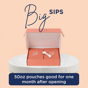 Open Milk by Mom shipping box containing two large pouches and a two in one scoop, displayed on a round marble stand. Text above reads 'Big Sips,' and a banner below says '50oz pouches good for one month after opening.