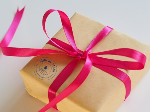 Gift box wrapped in brown paper with a bright pink ribbon bow, featuring the Milk by Mom logo.