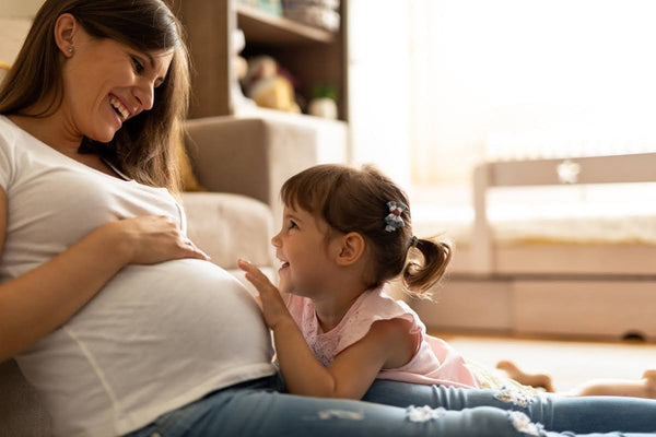 Smiling pregnant woman sitting on the floor with her young daughter touching her baby bump