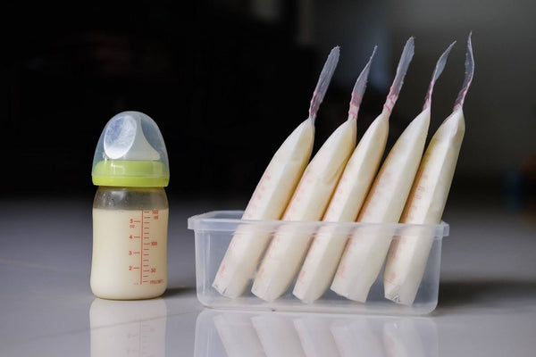 Baby bottle filled with milk placed beside five breast milk storage bags in a plastic container.