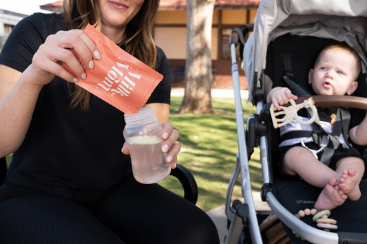 Mother pouring breast milk from a Milk MoM storage bag into a baby bottle while sitting outdoors, with her baby in a stroller watching