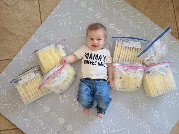 Smiling baby in a 'Mama’s Coffee Date' shirt lying on a mat surrounded by large freezer bags filled with stored breast milk