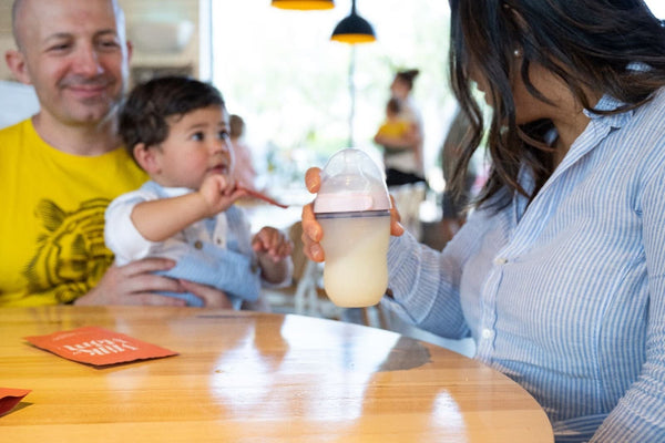 Mother holding a baby bottle of milk at a table while father holds their young child reaching toward the bottle