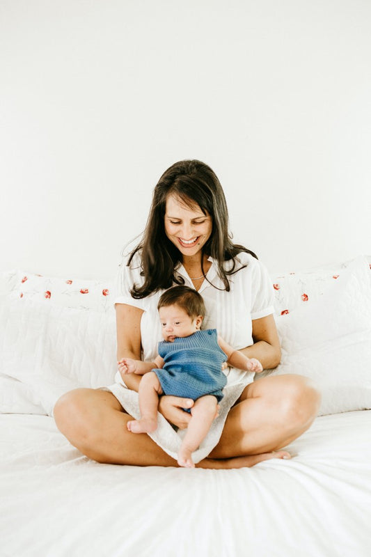Mother sitting cross-legged on a bed, smiling while holding her baby dressed in a blue outfit