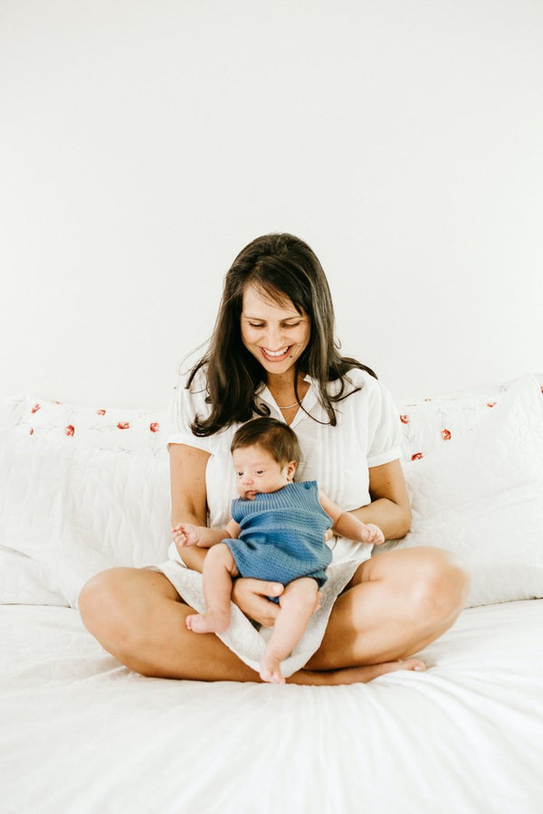 Mother sitting cross-legged on a bed, smiling while holding her baby dressed in a blue outfit