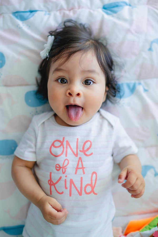 Baby lying on a blanket with whale patterns, sticking out their tongue and wearing a white onesie that says 'One of a Kind' in red letters