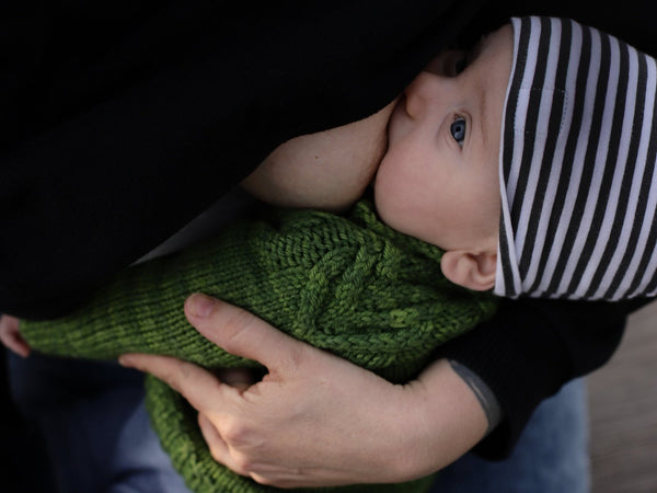 Baby in a green knitted outfit and striped hat breastfeeding while being held in her mom’s arms.