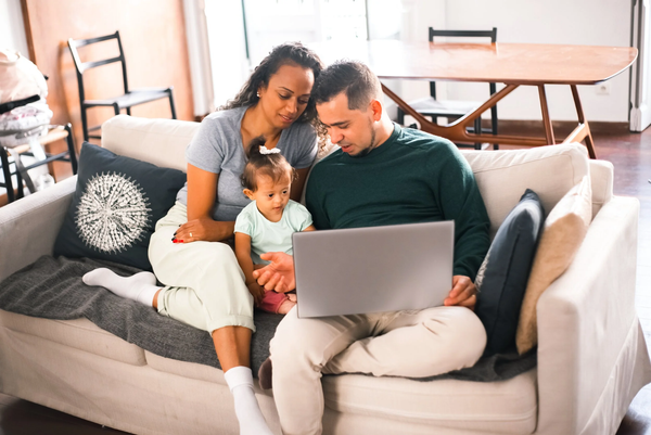 Family sitting on a couch together with a toddler, looking at a laptop screen.