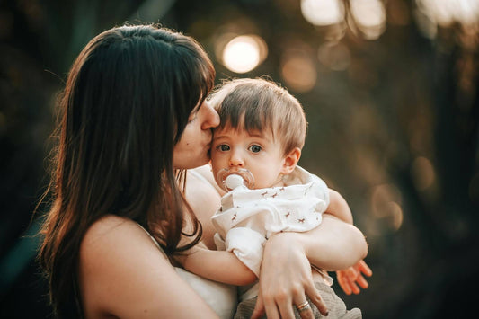 Mother kissing her baby on the cheek while holding him outdoors at sunset, tender parenting moment showing love, bonding, and family connection.