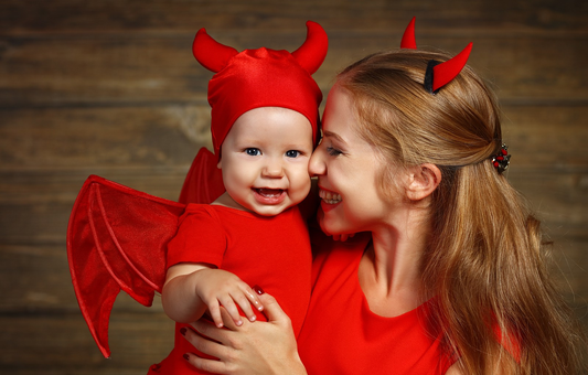Smiling mother and baby dressed in matching red devil costumes with horns, wings, and red outfits