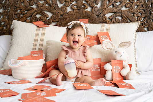 Smiling baby sitting on a bed surrounded by Milk by Mom freeze-dried breast milk pouches, holding a bottle of rehydrated breast milk, showcasing convenience, nutrition, and family-friendly feeding solutions