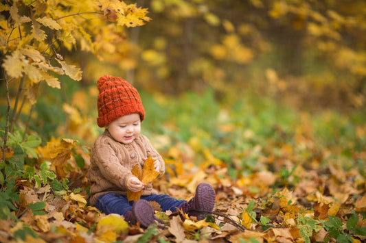 Baby wearing a red knitted hat and brown sweater and blue jeans sitting on autumn leaves while holding a yellow leaf.