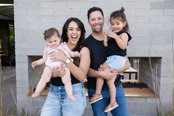 Smiling family of four standing outdoors, parents holding two young daughters while posing for a photo.