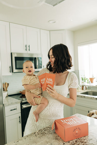 Mom holding baby in kitchen with Milk by Mom freeze-dried breast milk pouches and shipping box, highlighting convenient breast milk preservation and easy feeding solutions for busy families