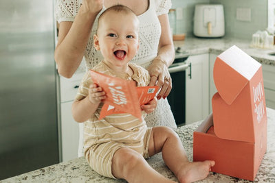 Baby holding Milk by Mom freeze-dried breast milk pouch next to packaging box in kitchen, shelf-stable breast milk solution for convenient baby feeding and milk storage