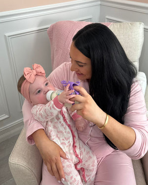 Mother sitting in a chair feeding her baby with a bottle while using a bottle toy attachment to keep the baby engaged during feeding time