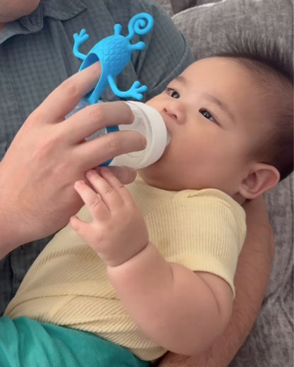 Baby being bottle-fed while holding a blue silicone bottle toy attachment that helps keep the baby engaged and focused during feeding