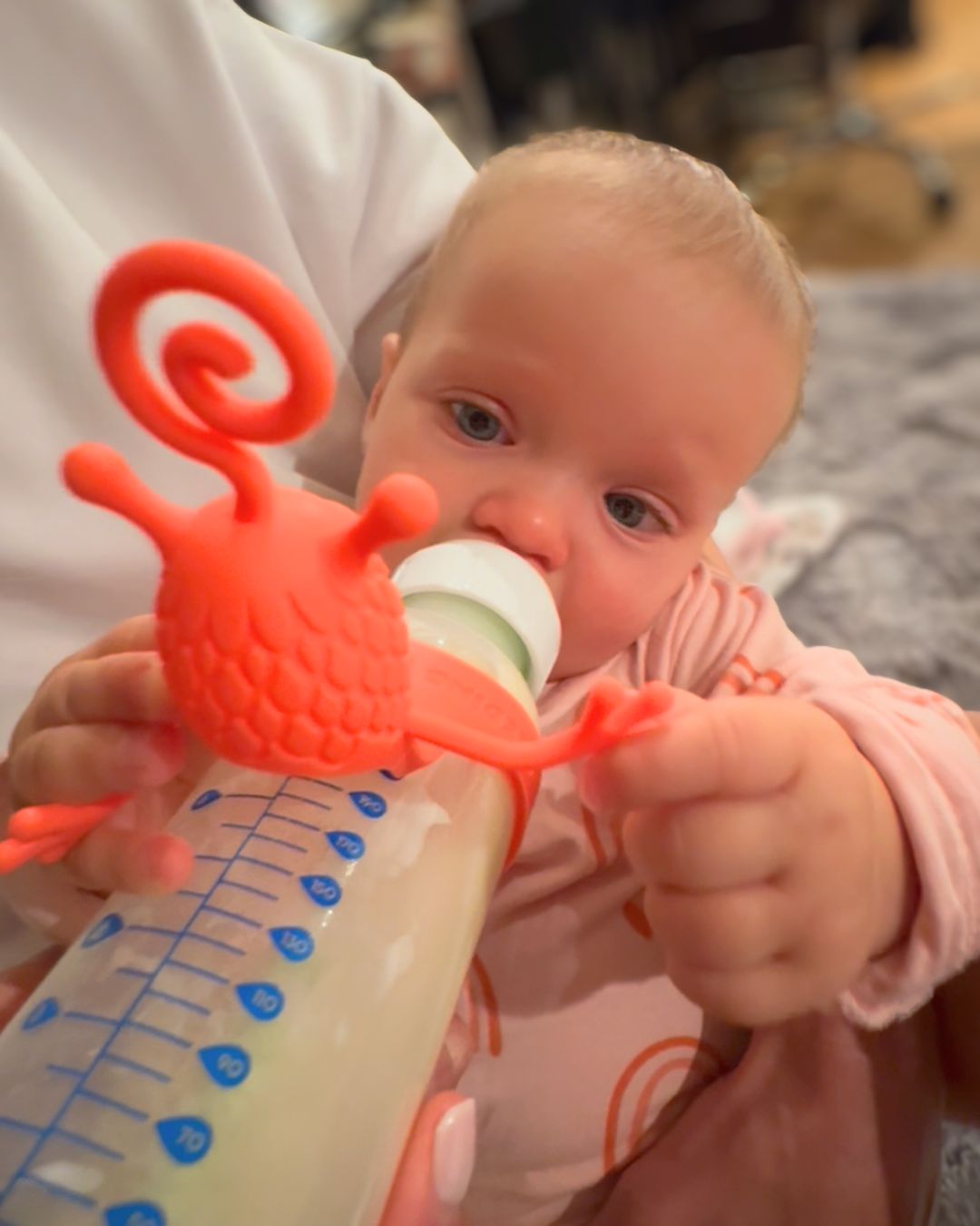Baby drinking from a bottle while holding a bright red silicone bottle toy attachment that helps keep the baby engaged and focused during feeding