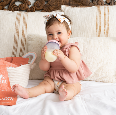 Smiling baby sitting on a bed and enjoying a bottle of reconstituted Milk by Mom freeze-dried breast milk, with pouches nearby, highlighting the ease and convenience of feeding with breast milk