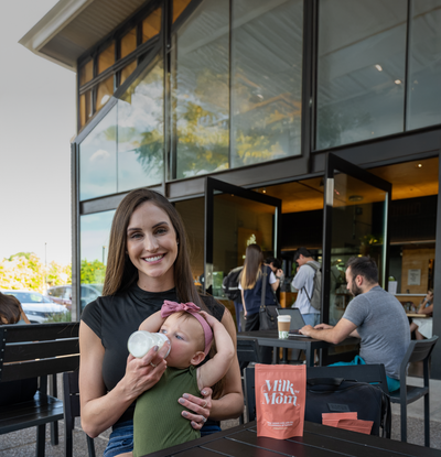 Mom feeding her baby a bottle at an outdoor café with Milk by Mom freeze-dried breast milk packets on the table