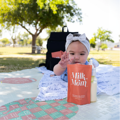 Baby reaching for a Milk by Mom freeze-dried breast milk pouch while lying on a blanket outdoors, showcasing the convenience and portability of freeze-dried breast milk for on-the-go families