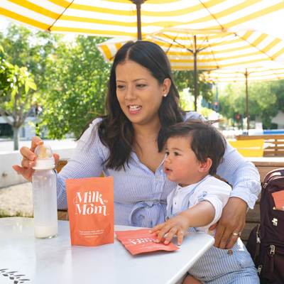 Mom preparing a bottle with Milk by Mom freeze-dried breast milk pouch while sitting outdoors with her toddler, highlighting easy mixing, portable breast milk nutrition, and convenient feeding on the go